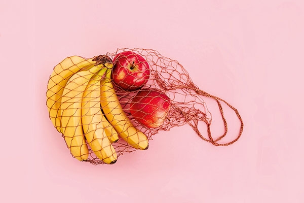 Reusable bag with apples and bananas on pink background. Zero waste and plastic free shopping concept.